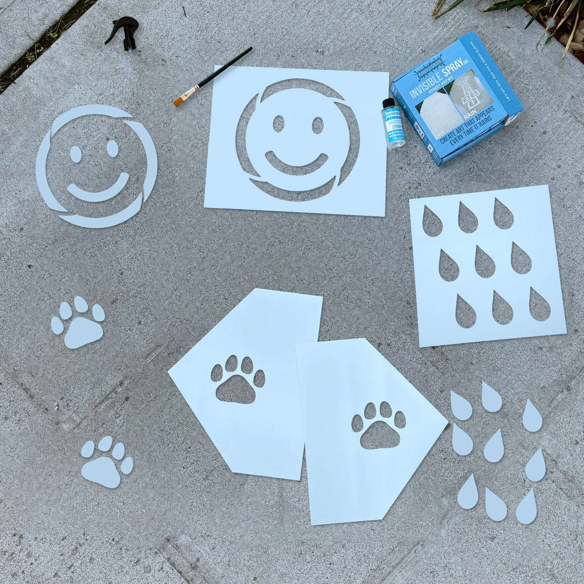 The Rainmaker Bundle laid out on a sidewalk: Invisible Spray kit, smiley face, paw print and raindrop stencils, and a paintbrush