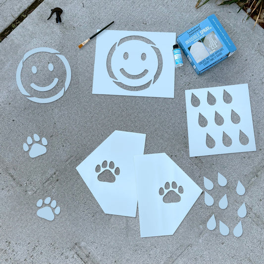 The Rainmaker Bundle laid out on a sidewalk: Invisible Spray kit, smiley face, paw print and raindrop stencils, and a paintbrush